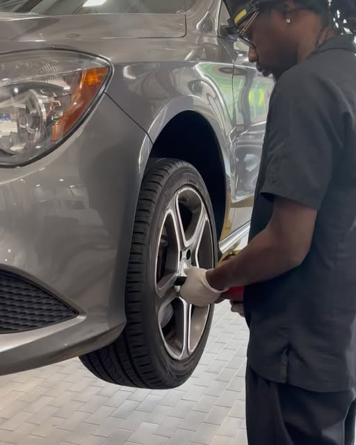 A Suivels Auto mechanic crouched at the front wheel of a silver sedan on a paved driveway.